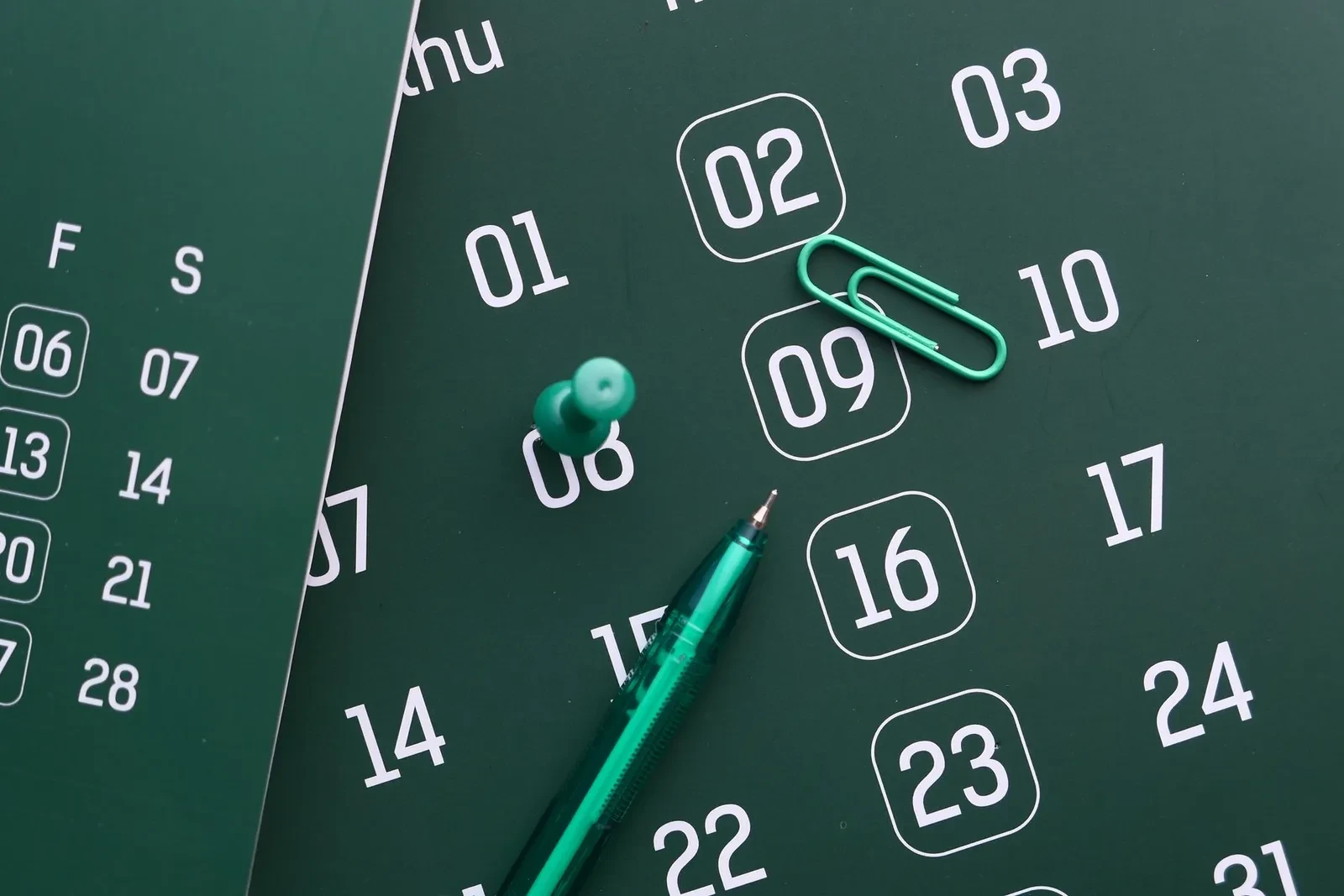 Un calendario verde con varias fechas marcadas, una chincheta y un bolígrafo