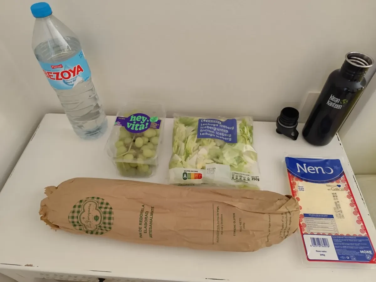 Pan, queso en lonchas, ensalada, uvas y agua sobre una mesa blanca