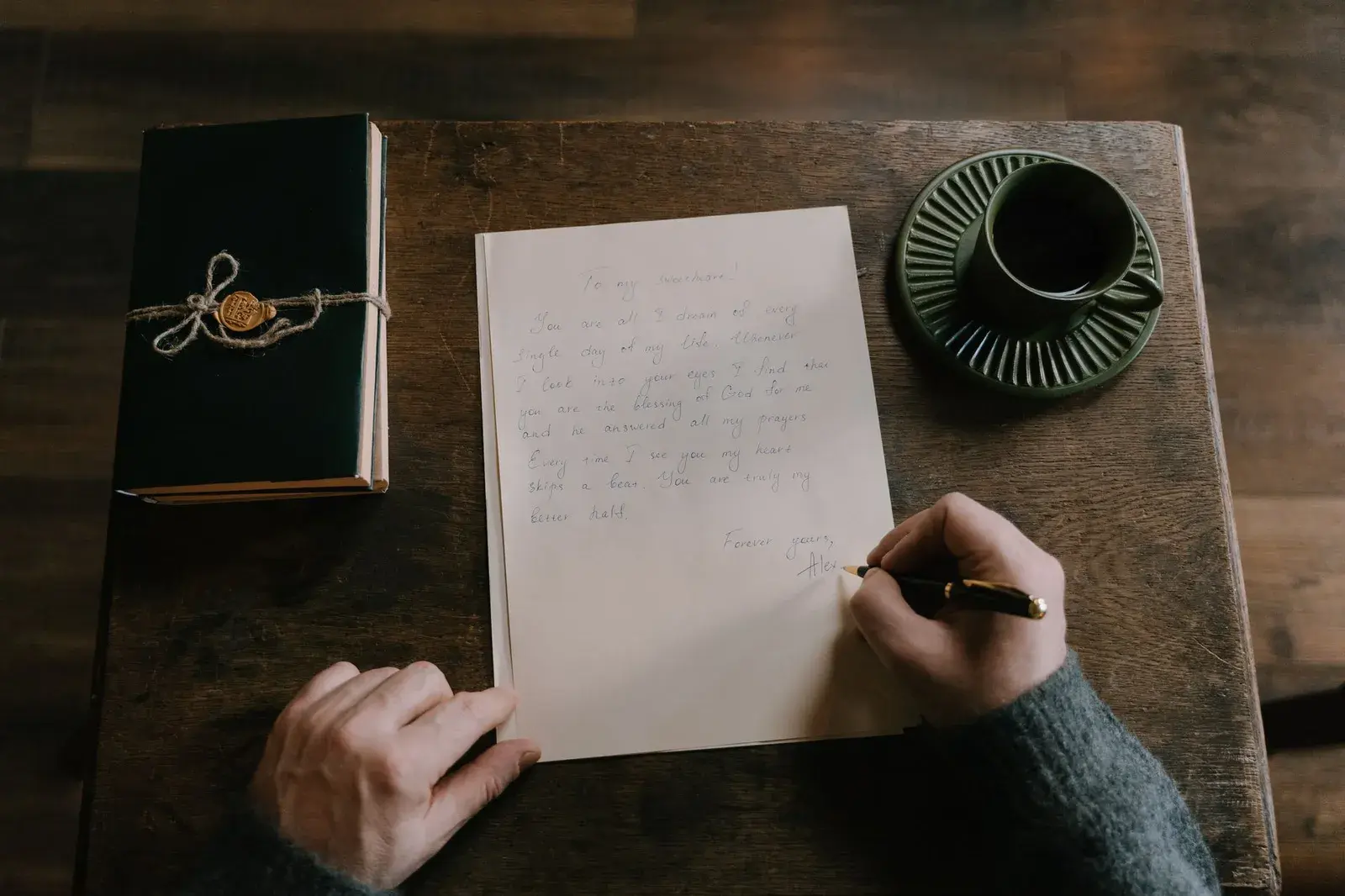Una persona escribiendo una carta a mano en una mesa de madera junto a una libreta y un café