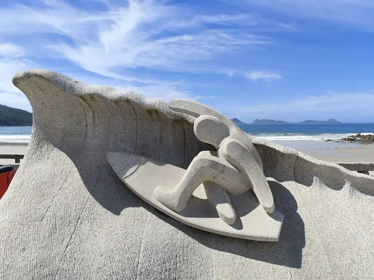 Escultura de piedra de un surfista sobre una ola, con las islas Cíes al fondo