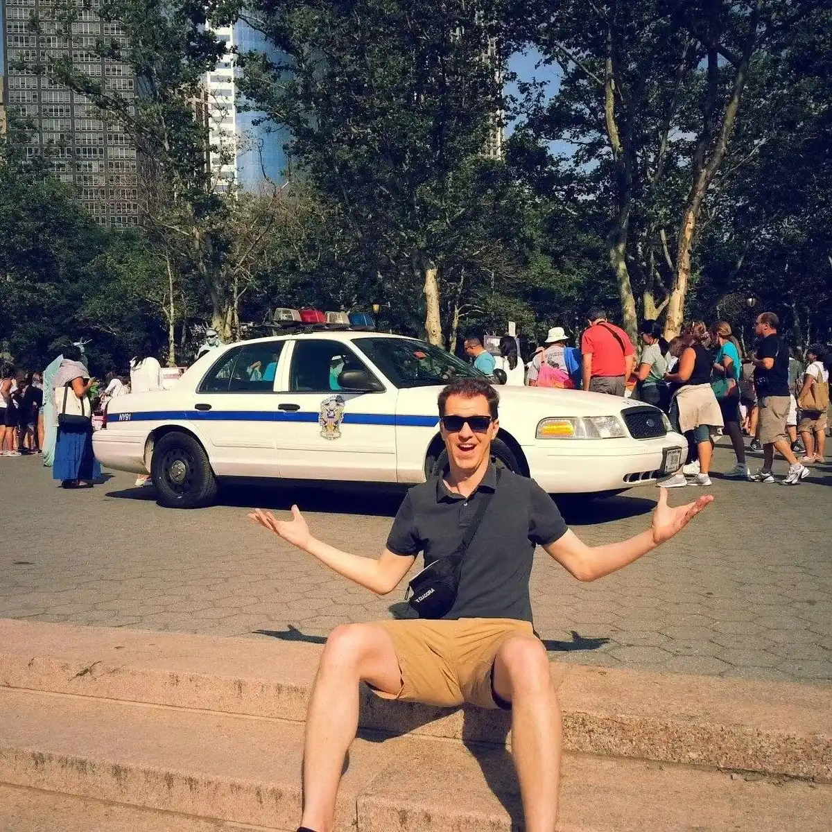 Michiel Das sentado en unos escalones de Battery Park con los brazos abiertos, con un coche de policía y turistas detrás